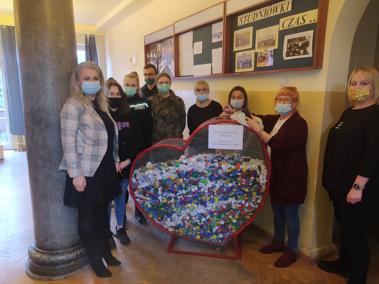 Młodzież wraz z dyrekcją szkoły stoi przy metalowym sercu wypełnionym nakrętkami