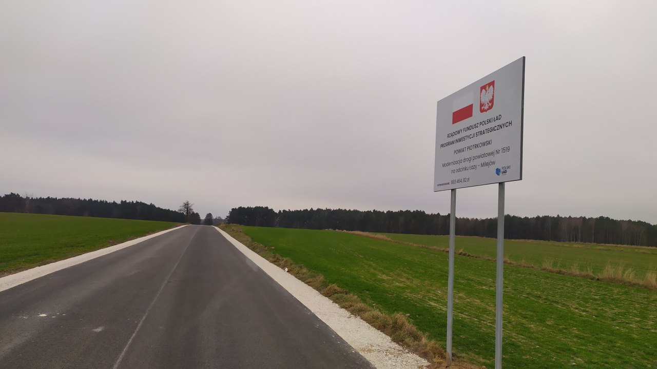 Fragment drogi powiatowej Łazy - Milejów. Po prawej stronie tablica informująca o otrzymanym dofinansowaniu.