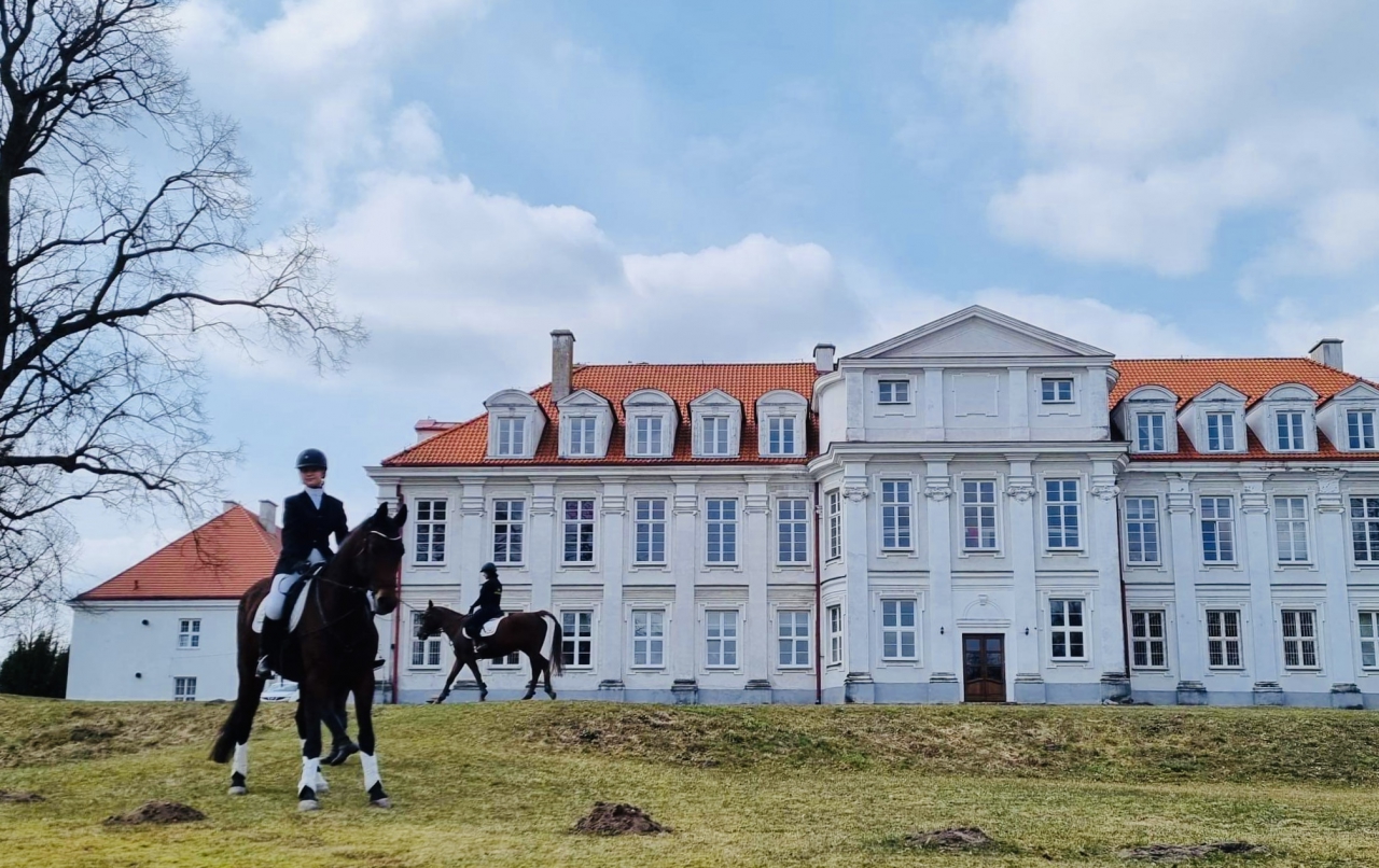 Budynek szkoły w Wolborzu z zewnątrz. Przed budynkiem dwie osoby jeżdżą na koniach.