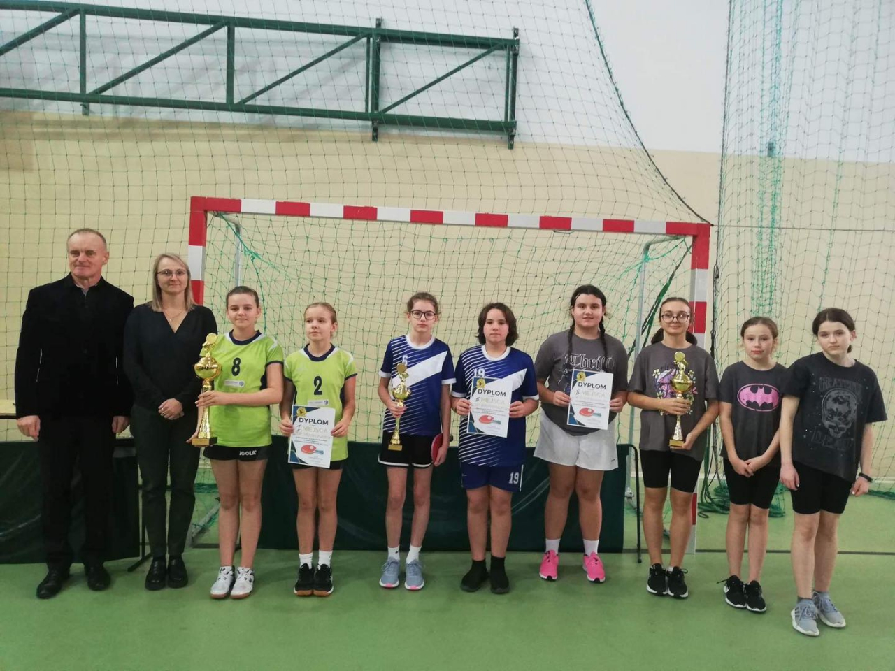 Zwycięskie drużyny dziewcząt stoją w hali sportowej z pucharami
