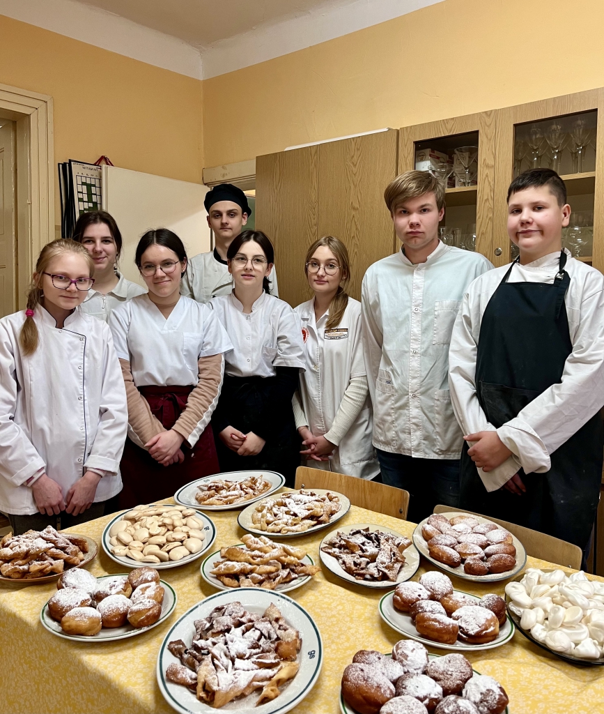 Stół ze słodkimi pączkami, faworkami i bezami. W tle uczniowie klasy w zawodzie technik żywienia i usług gastronomicznych, ściana i meble