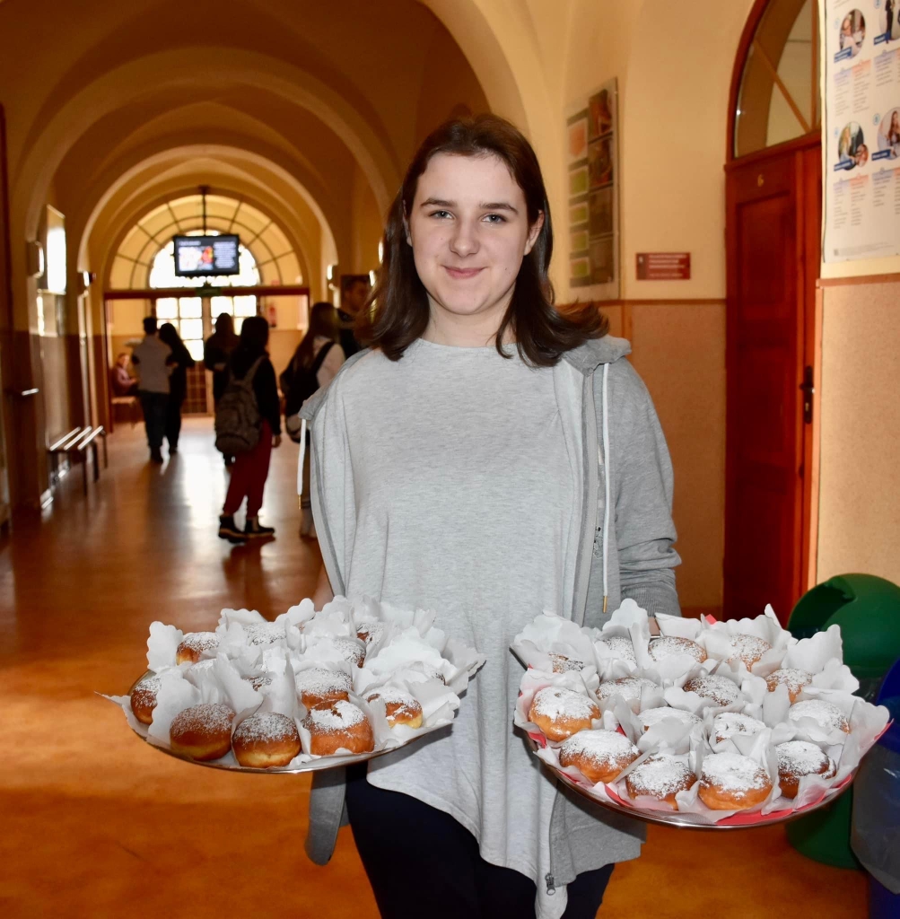 Uczennica z tacami pełnymi pączków. W tle hol szkoły, drzwi do sal, w oddali okno i inni uczniowie.