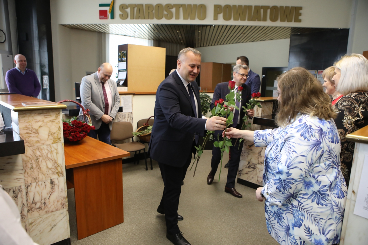 Starosta Piotr Wojtysiak wraz z władzami Powiatu znajduje się w pomieszczeniu podczas wręczania kwiatów pracownicom. w tle napis Powiat Piotrkowski