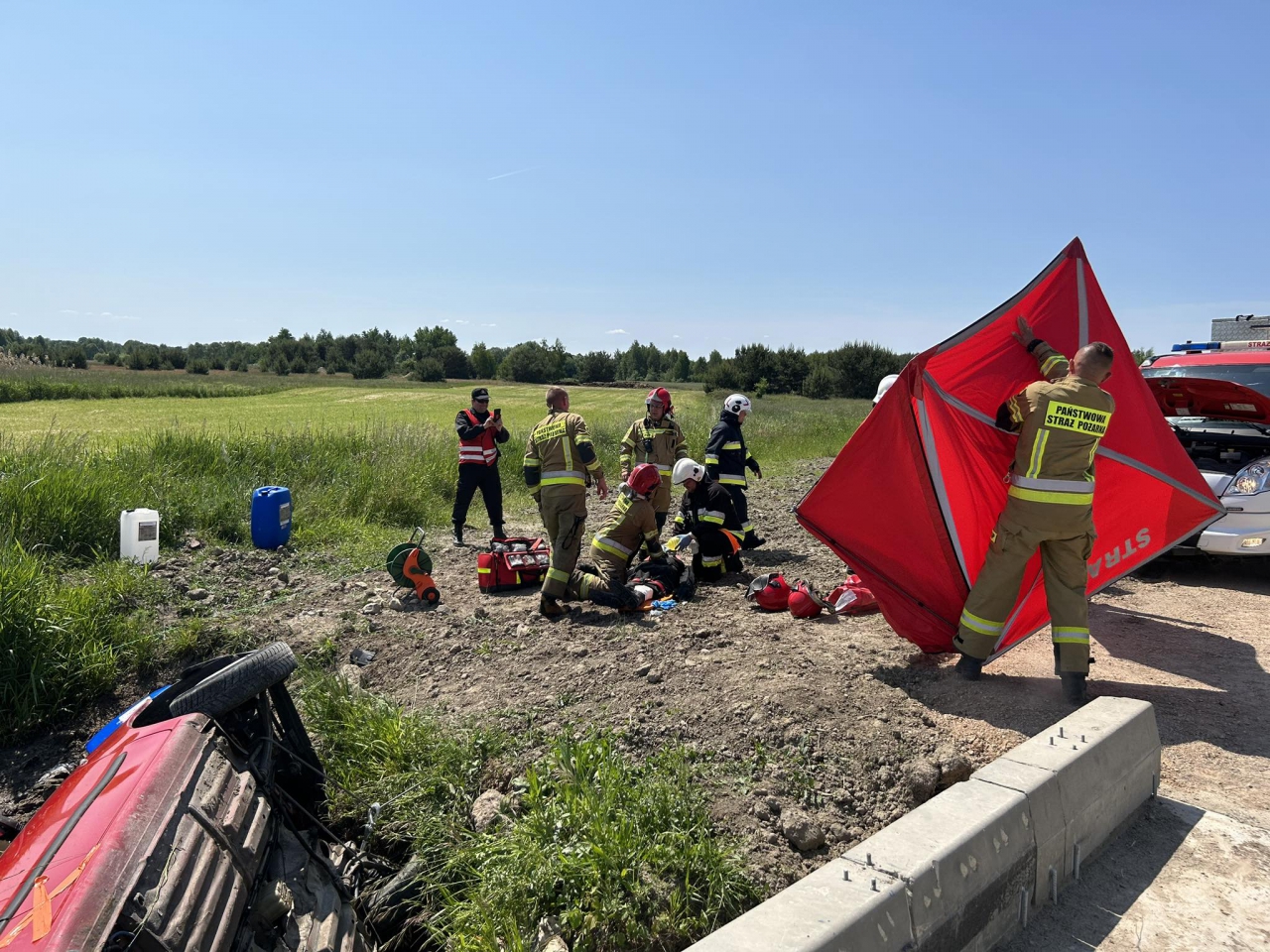 Strażacy udzielają pomocy rannemu, w rzece leży przewrócone auto