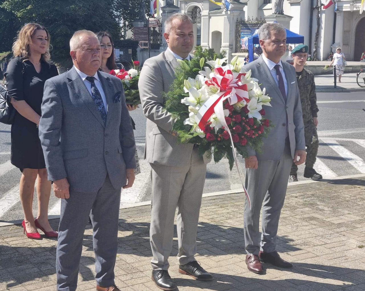 Władze Powiatu Piotrkowskiego podczas składania kwiatów stoją na świeżym powietrzu.